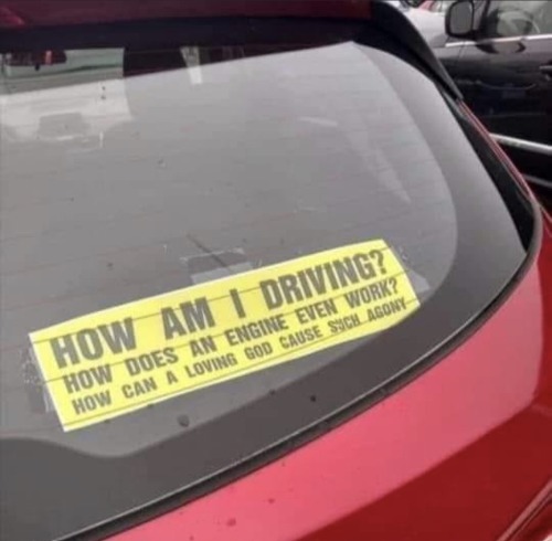 A yellow bumpersticker in the rear window of a red car: “HOW AM I DRIVING? HOW DOES AN ENGINE EVEN WORK? HOW CAN A LOVING GOD CAUSE SUCH AGONY”