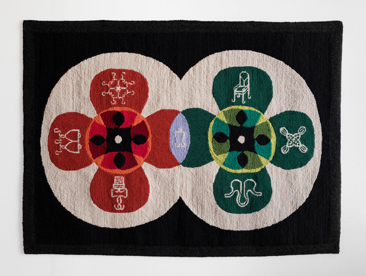 A textured rug with a black background and two intersecting white circles. Inside each circle four-lobed flower patterns (one green, one red) display a variety of symbols.