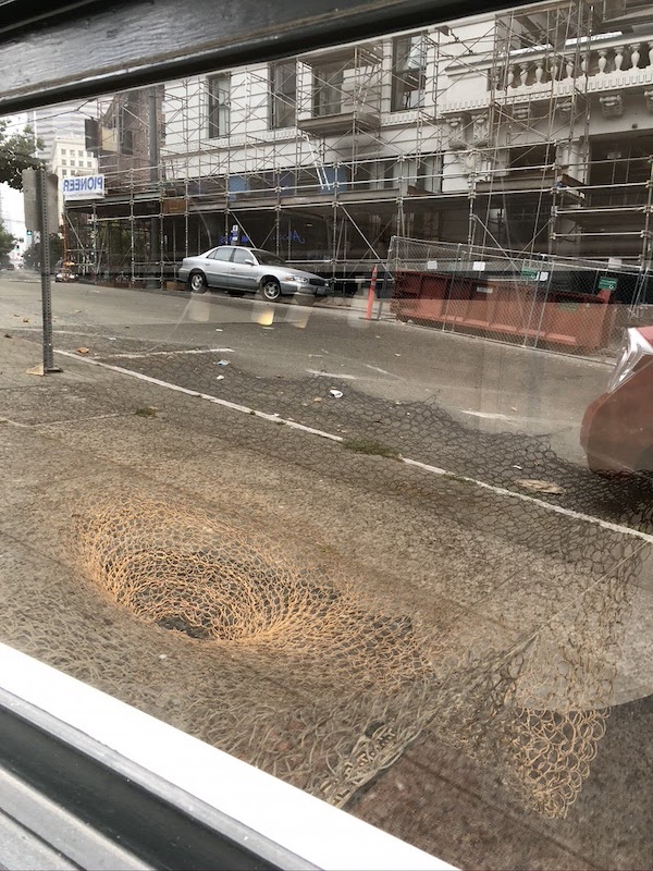 A window at street level presents a view into the gallery below. Hanako O’Leary’s Yomi is seen from above, jute netting woven down into a funnel. Reflected in the window are the city street, a construction site, a parked car, and a sign that reads “Pioneer” backwards.